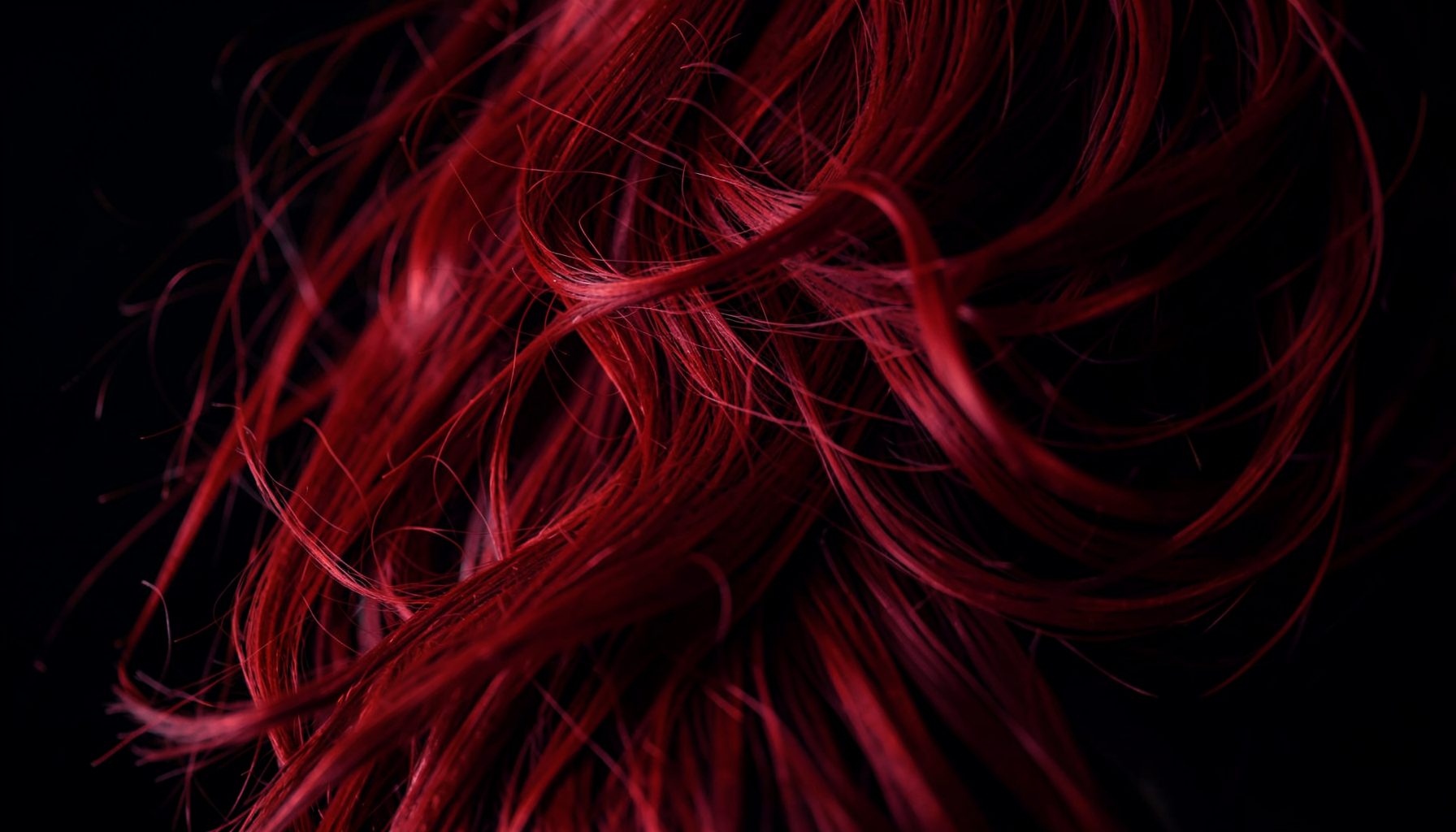 A close-up of red hair flowing against a dark background, subtle crimson highlights suggesting danger and mystery